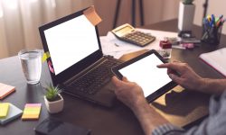 Mockup image of male hands using a laptop computer and holing a smart phone both with blank white screens. Focus on the index finger